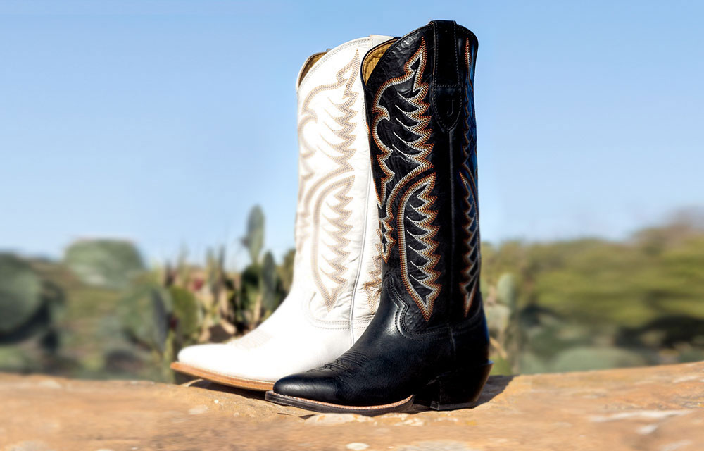 The Evelyn 15&rdquo; Western Boot in colors of coal and vintage ivory sitting on a rock with cactus in the background. 